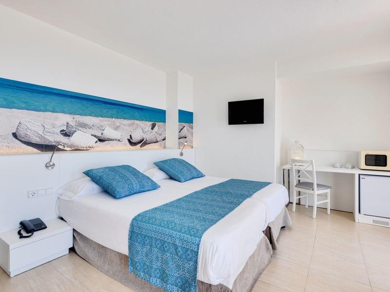 Modernes Schlafzimmer mit Doppelbett, blauer Bettdecke, zwei Kissen, TV, Schreibtisch und Strandbild.