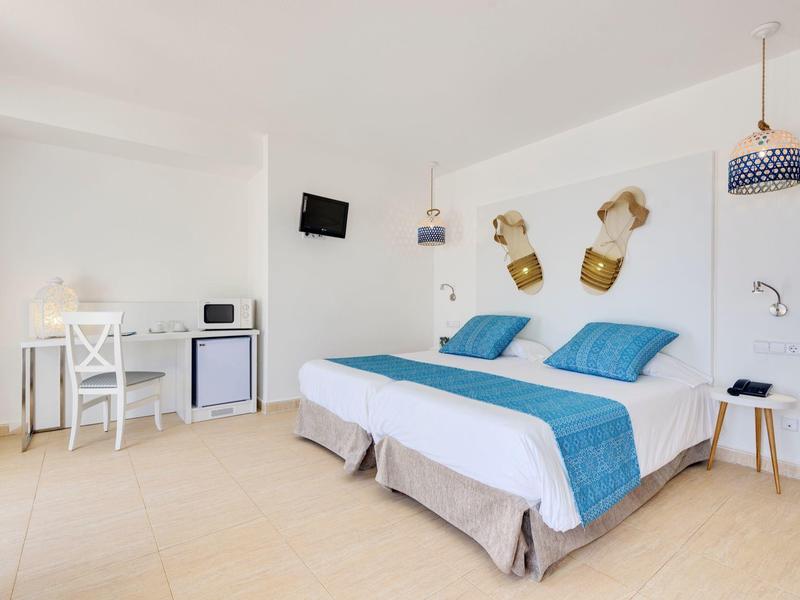 Helles Schlafzimmer mit Doppelbett, weiß-blauer Bettwäsche, Schreibtisch, Minikühlschrank und Wand-TV.