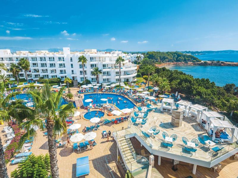 Hotelanlage mit Pool, Sonnenschirmen, Palmen und Blick auf das blaue Meer unter blauem Himmel.