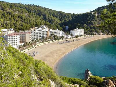 Klarer Strand mit goldenem Sand, umgeben von grünen Hügeln und mehreren weißen Gebäuden am Ufer.