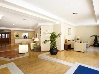 Heller Hotellobby mit weißen Sesseln, großen Pflanzen und heller Holz- und Fliesenboden.