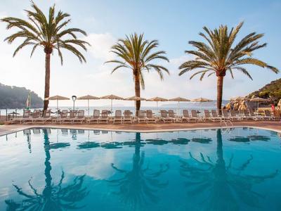 Schwimmbecken mit klarem Wasser, drei Palmen und weißen Sonnenliegen unter Sonnenschirmen.