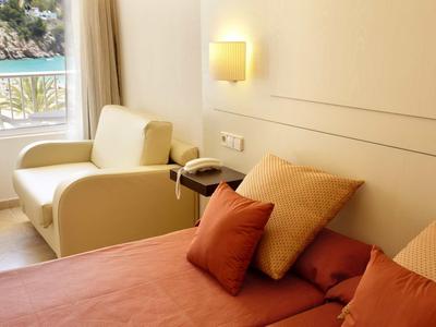 Gemütliches Schlafzimmer mit rotem Bett, Kissen und weißem Sessel neben Fenster und Vorhängen.