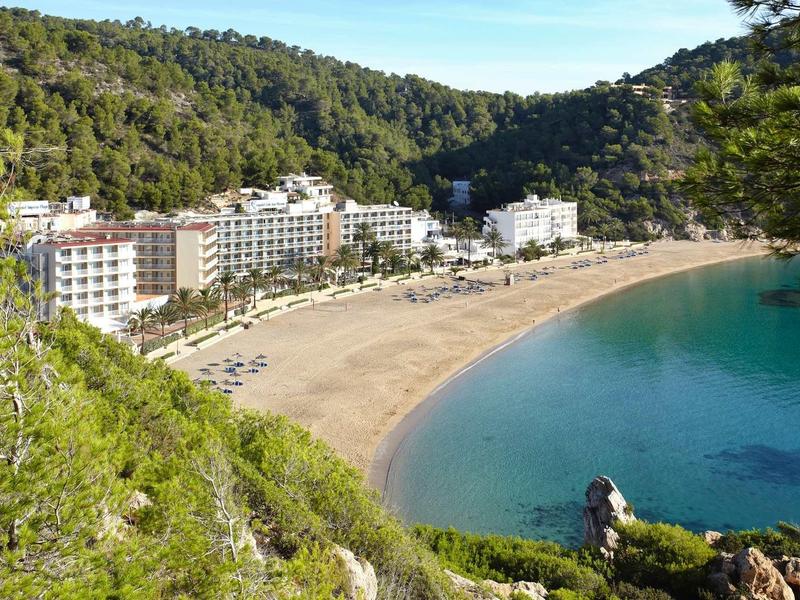 Klarer Strand mit goldenem Sand, umgeben von grünen Hügeln und mehreren weißen Gebäuden am Ufer.
