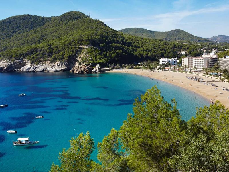 Klarer blauer Meer mit Booten, Sandstrand, bewaldete Hügel und Gebäude am Küstenrand.