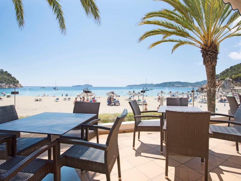 Leerer Sitzbereich mit Tischen und Stühlen am Strand mit Palmen und blauem Himmel im Hintergrund.
