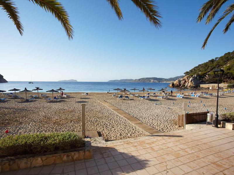 Strand mit Sand, Liegen und Sonnenschirmen, umgeben von Palmen und Hügeln am ruhigen Meer.