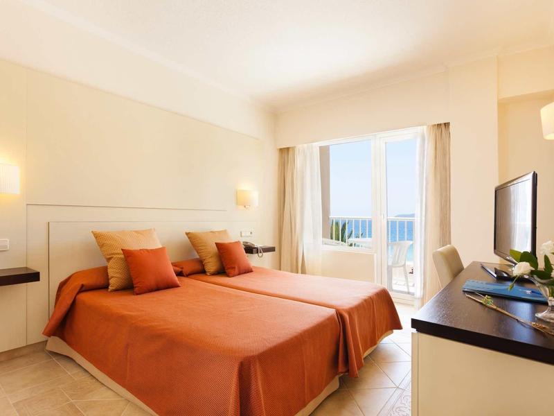 Helles Schlafzimmer mit zwei Betten, orangen Bettdecken und Balkon mit Meerblick.