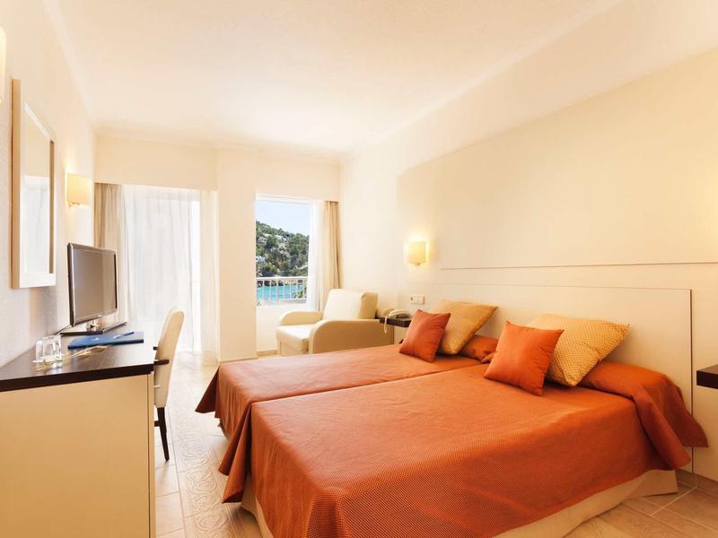 Helles Schlafzimmer mit zwei Betten, orangefarbenen Decken und Balkon mit Blick ins Grüne.