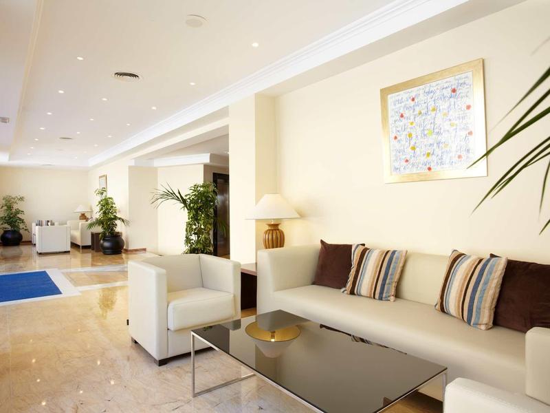 Heller Wohnzimmerbereich mit beige Wänden, grauen Sofas, Dekokissen und grünen Zimmerpflanzen.