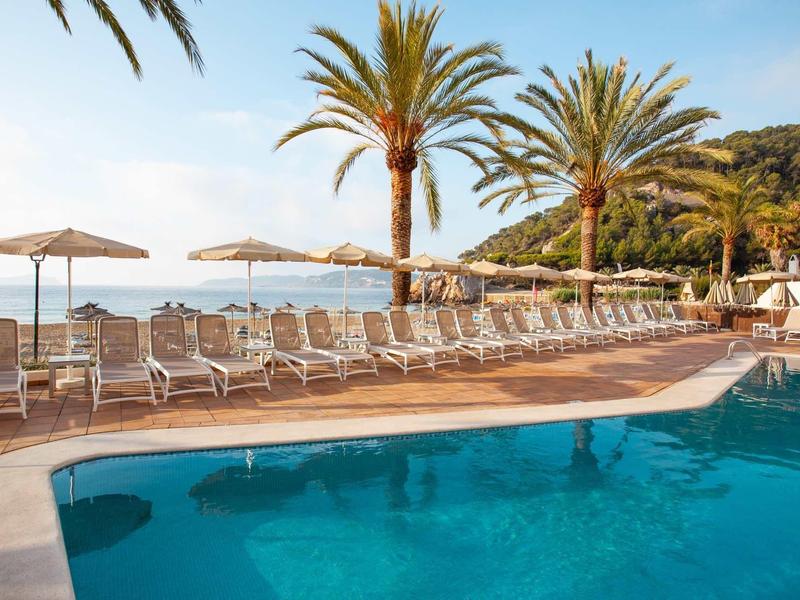 Einladender Pool mit klar blauem Wasser, Liegestühlen und Sonnenschirmen vor Palmen am Meer.