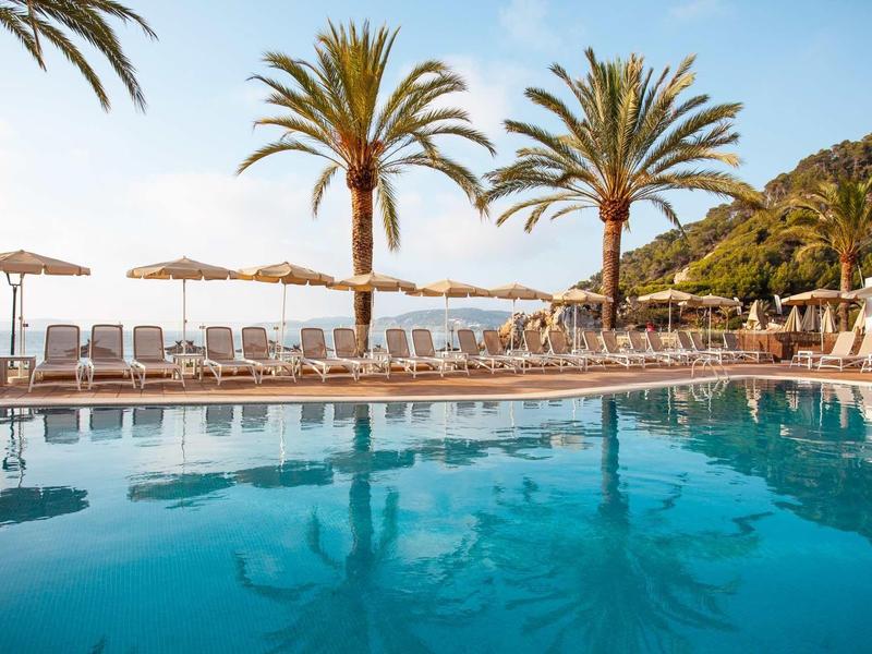 Blauer Pool mit Liegestühlen, Sonnenschirmen und Palmen unter klarem Himmel am Meer.