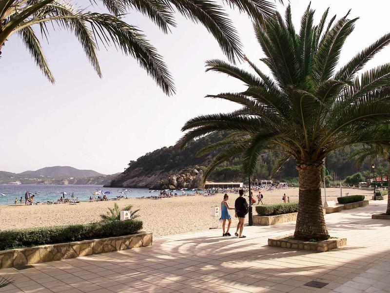 Strandpromenade mit Palmen, Spaziergängern, Sandstrand und ruhigem Meer im Hintergrund.
