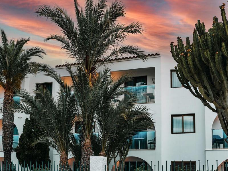 Biały budynek hotelowy z palmami i kaktusami o zachodzie słońca.