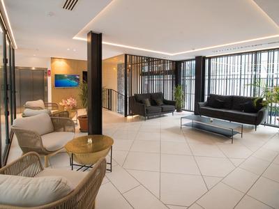 Heller, moderner Wartebereich mit weißen Kacheln, schwarzen Sofas, Stühlen, Tischen und großen Fenstern.