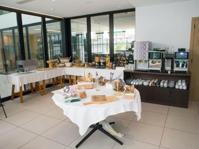 Ein heller Frühstücksraum mit weißen Tischen, Kaffeeautomaten und einem Buffet mit Snacks und Getränken.