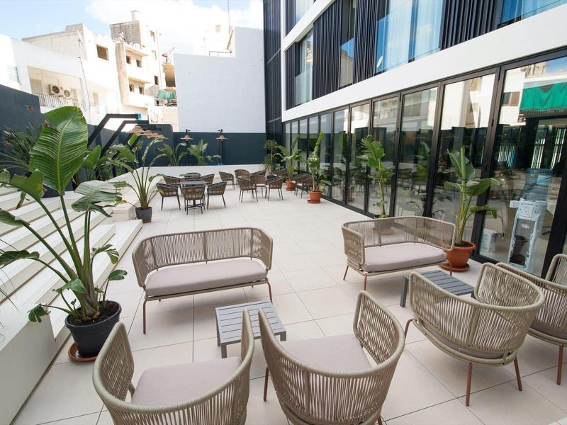 Moderner Außenbereich mit grauen Sesseln, Pflanzen und Glasfenstern an hellem Boden.