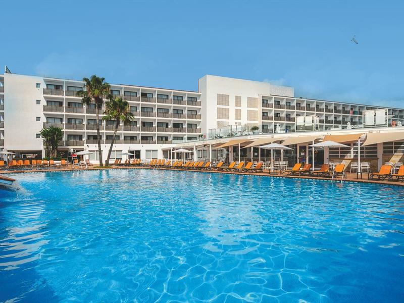 Groot hotelzwembad met ligstoelen en palmbomen voor een wit meerlaagse hotelgebouw onder een blauwe lucht