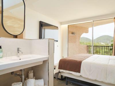 Habitación de hotel luminosa con cama, lavabo y vista a la montaña desde un gran balcón.