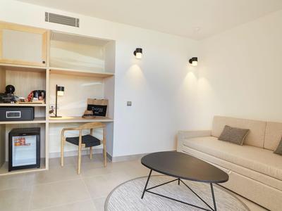 Habitación de hotel moderna con escritorio, mini nevera, estantes y sofá acogedor.