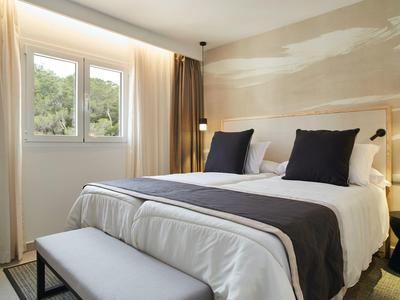 Dormitorio moderno y luminoso con cama grande, almohadas blancas y oscuras y ventana con vista a la vegetación.