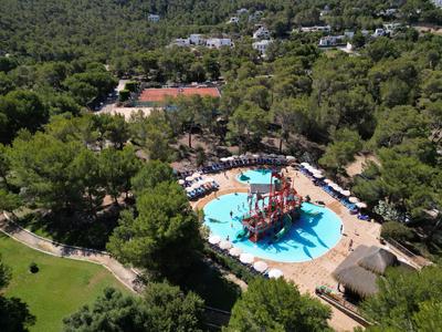 Parque acuático con barco pirata y toboganes, rodeado de árboles verdes y una pista de tenis.