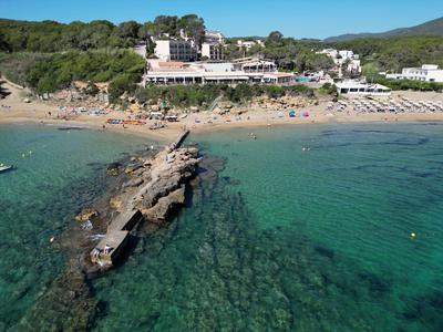 Agua clara con muelle rocoso y playa de arena concurrida junto a hoteles y colinas boscosas.