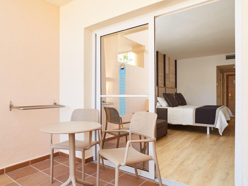 Balcón con mesa y sillas, dormitorio con cama doble visible a través de puerta corredera de vidrio