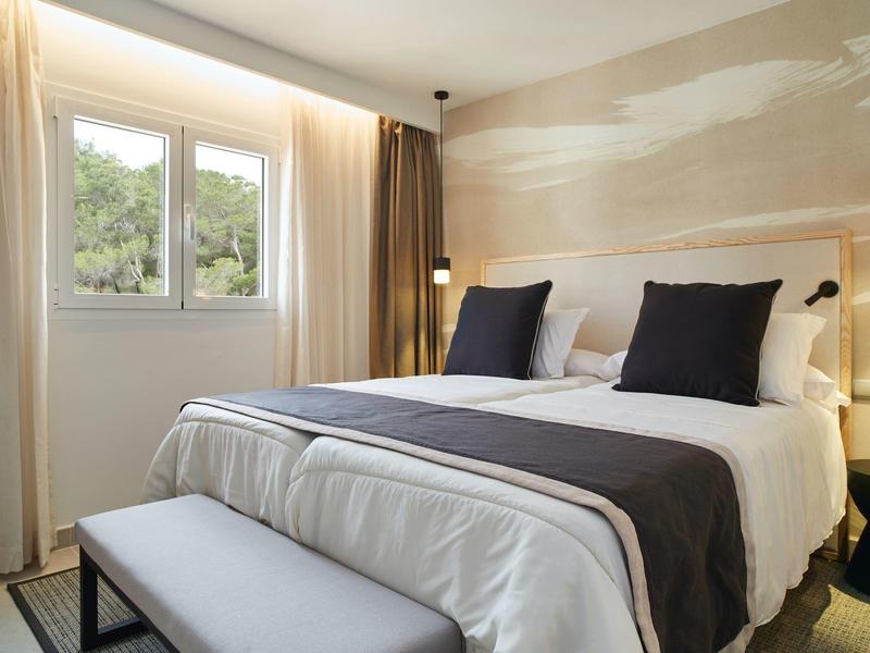 Dormitorio moderno y luminoso con cama grande, almohadas blancas y oscuras y ventana con vista a la vegetación.