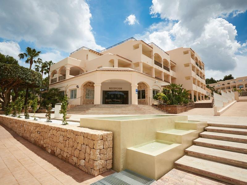 Hotel moderno con terraza, escaleras y palmeras bajo un cielo azul con nubes