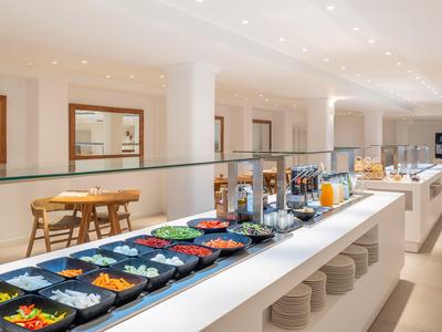 Área de desayuno luminosa con buffet, frutas y jugos en un hotel moderno.