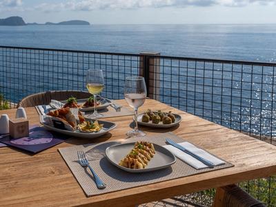 Mesa de madera con platos, bebidas y aperitivos en una terraza con vista al mar.