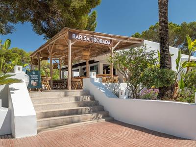 Área exterior de un bar con asientos y pabellón de madera sombreado, rodeado de vegetación mediterránea.