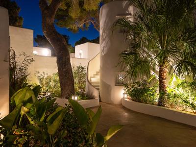 Camino de jardín iluminado con palmeras y vegetación junto a edificios blancos en un hotel.