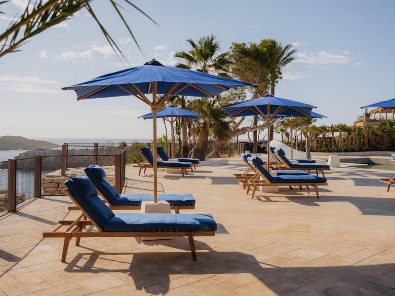 Leżaki z niebieskimi poduszkami pod niebieskimi parasolami na tarasie z widokiem na morze.