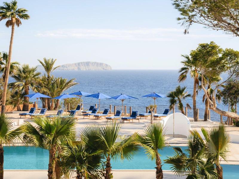 Basen hotelowy z leżakami i niebieskimi parasolami, z widokiem na morze i małą wyspę.