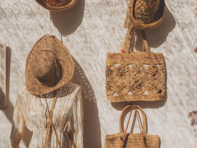 Ściana z dekoracyjnymi słomianymi kapeluszami, torbami i ubraniami w naturalnych kolorach.