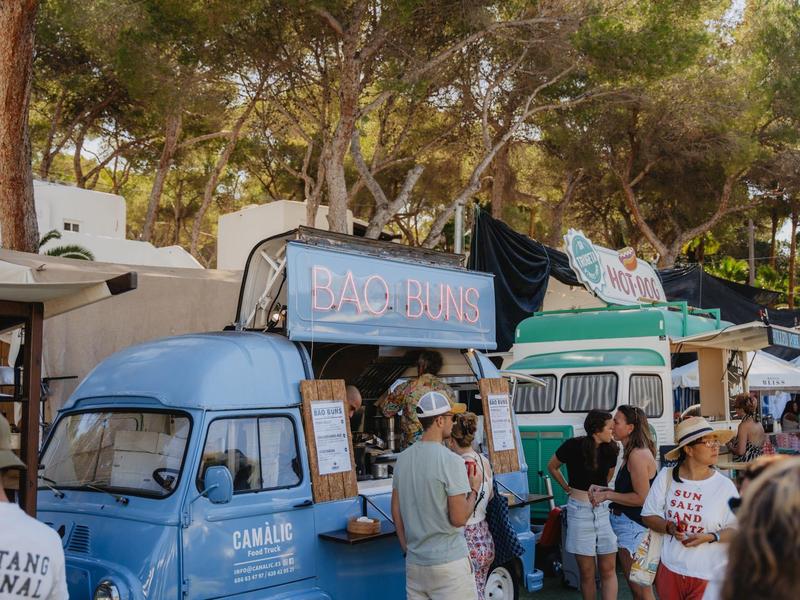 Ludzie gromadzą się przed różnymi niebieskimi food truckami na terenie z drzewami.