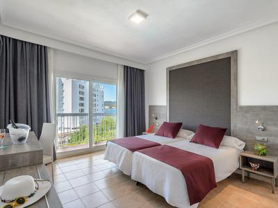 Jasny pokój hotelowy z dwoma łóżkami, balkonem i nowoczesnym umeblowaniem w neutralnych kolorach.