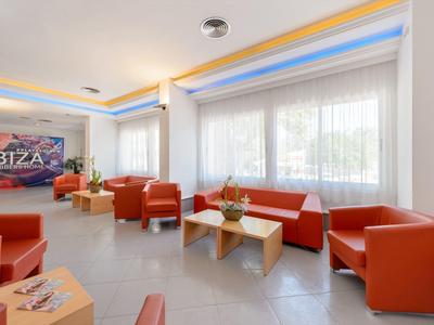 Jasny salon hotelowy z czerwonymi fotelami, sofą i stolikami przed dużymi oknami z białymi zasłonami.