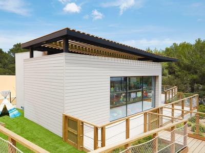 Modern klein huis met grote glazen voorkant en houten balustrade, omgeven door bomen.