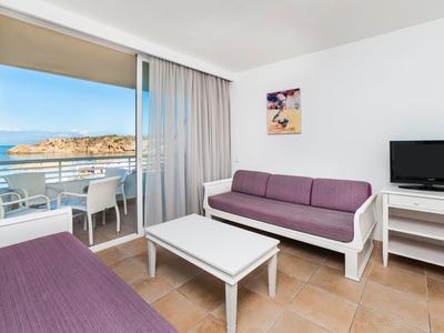 Licht hotelkamer met paarse banken, tv en balkon met uitzicht op zee.