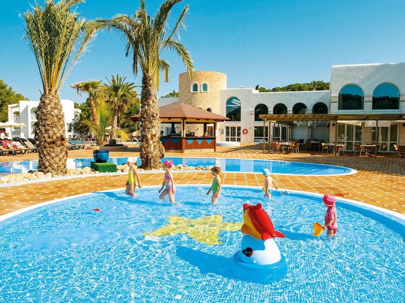 Kinderen spelen in een ondiep, rond hotelzwembad met palmbomen en ligbedden.