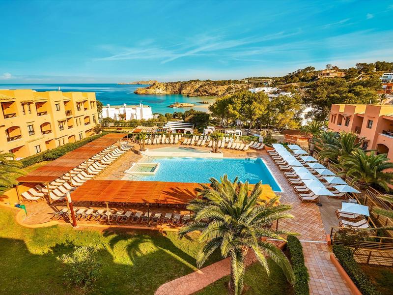 Hotelcomplex met zwembad, talloze ligstoelen, palmbomen en uitzicht op zee en kustlijn.