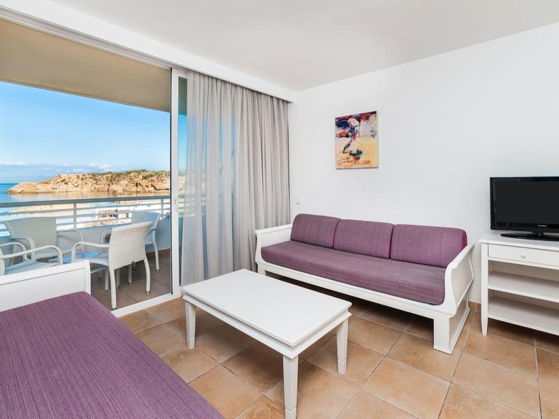 Licht hotelkamer met paarse banken, tv en balkon met uitzicht op zee.