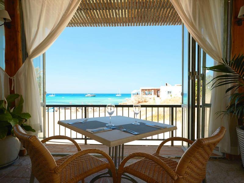 Uitzicht door open gordijnen op balkon met tafel en stoelen, zee en gebouwen daarachter.