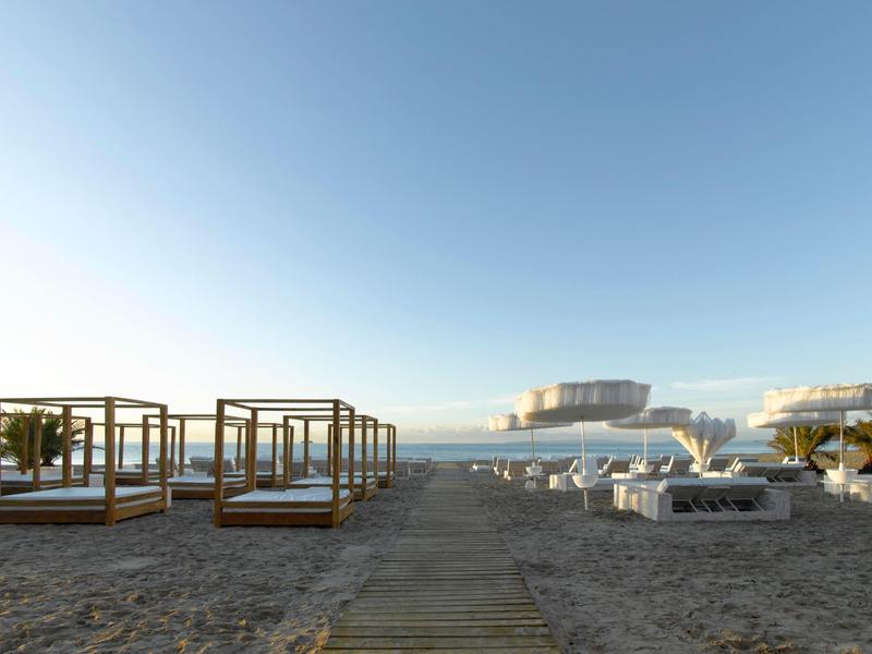 Plaża z białymi leżakami i parasolami, drewniane pawilony, widok na morze.