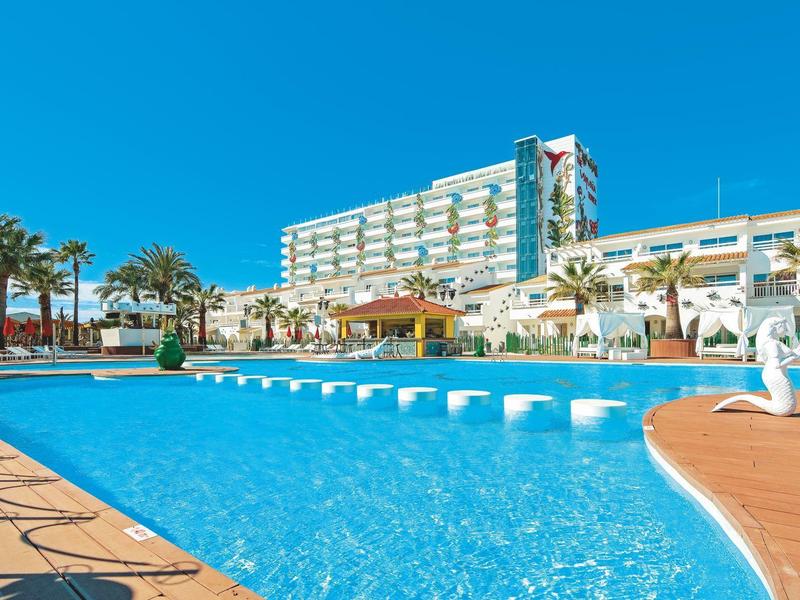 Przestronny basen na zewnątrz z nowoczesnym hotelem i palmami pod jasnym niebem.