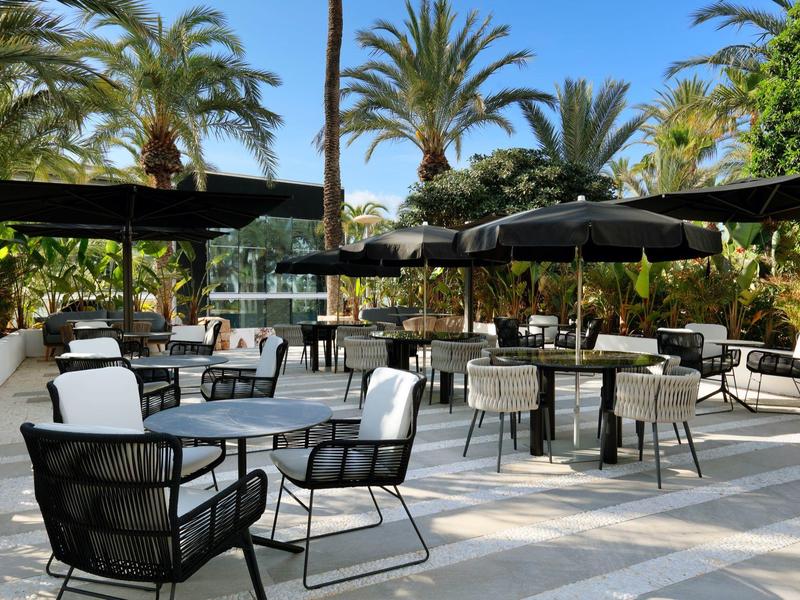 Zewnętrzny taras z stolikami i krzesłami pod parasolami, otoczony palmami.