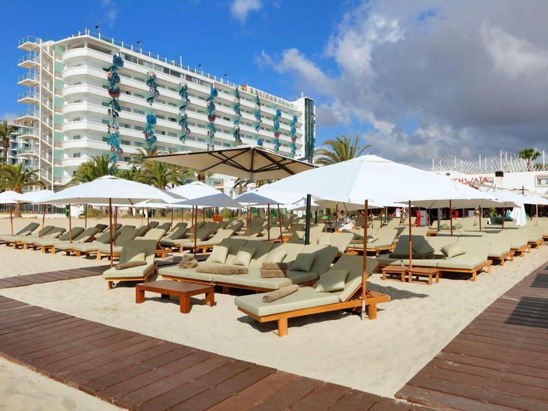 Piaszczysta plaża z leżakami i parasolami przed ośmiopiętrowym hotelem pod niebieskim niebem.